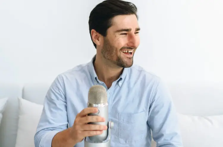 Man smiling while holding a Blue Yeti microphone, preparing to record the perfect podcast in a bright, minimal setup.