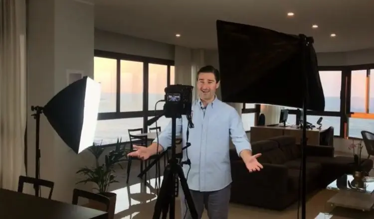 Liam Austin standing in front of a camera and softbox lights, recording a video podcast from his home setup with a scenic ocean view in the background