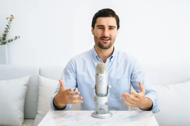 Man sitting at a table with a microphone, gesturing as he speaks during a podcast host recording session.