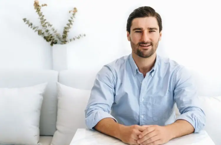 Liam Austin sitting at a table in a light blue button-down shirt, demonstrating the likely attire for male guest speaker appearances.