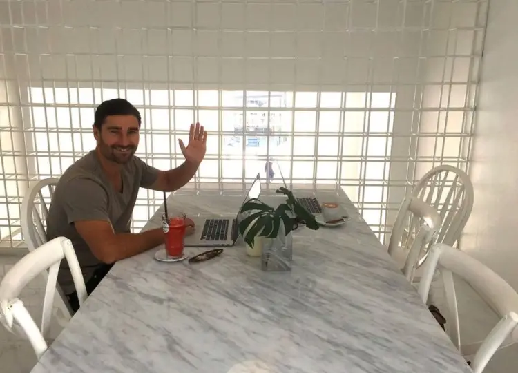 Liam Austin sitting at a marble table with his laptop and a drink, waving while working remotely, showing a casual setup someone might use when preparing for a great interview.