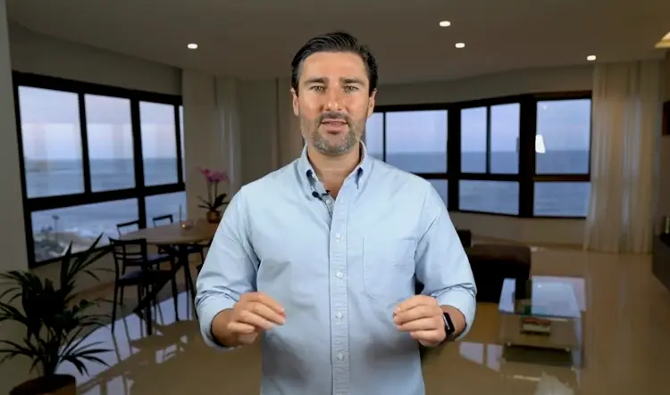 A man in a light blue shirt demonstrating presentation skills while speaking directly to the camera in a bright room.